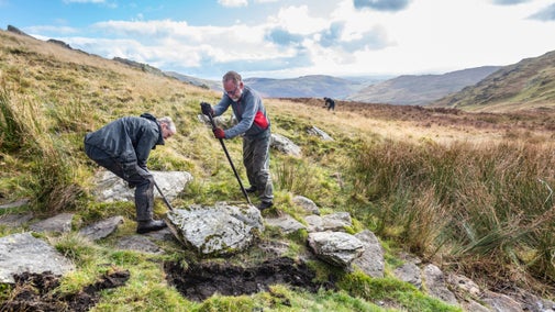 Three people using tools to moves stones from a grassy cliff landscape
